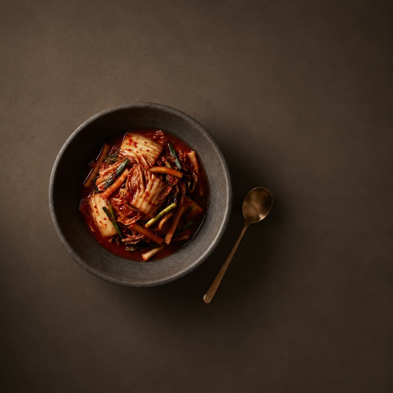 A bowl of house kimchi with a brass spoon on warm dark stone
